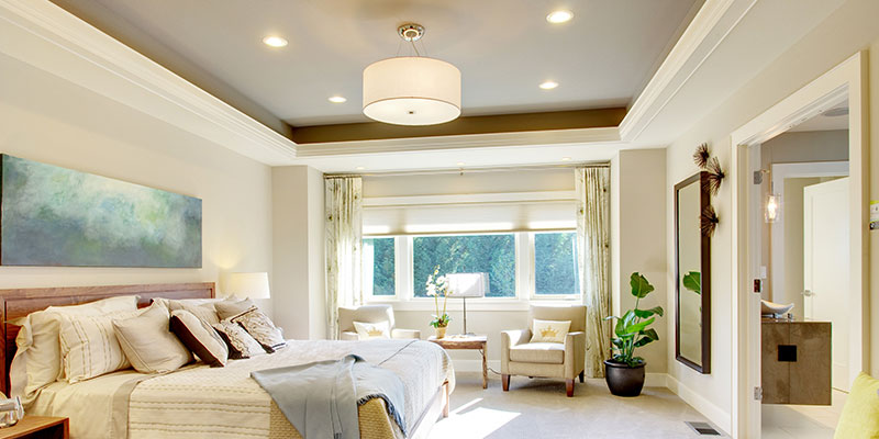 Bright, neutral-toned primary bedroom featuring layered lighting (recessed cans, drum pendant, bedside lamps), motorized roller shades, and a seating nook by a large window.