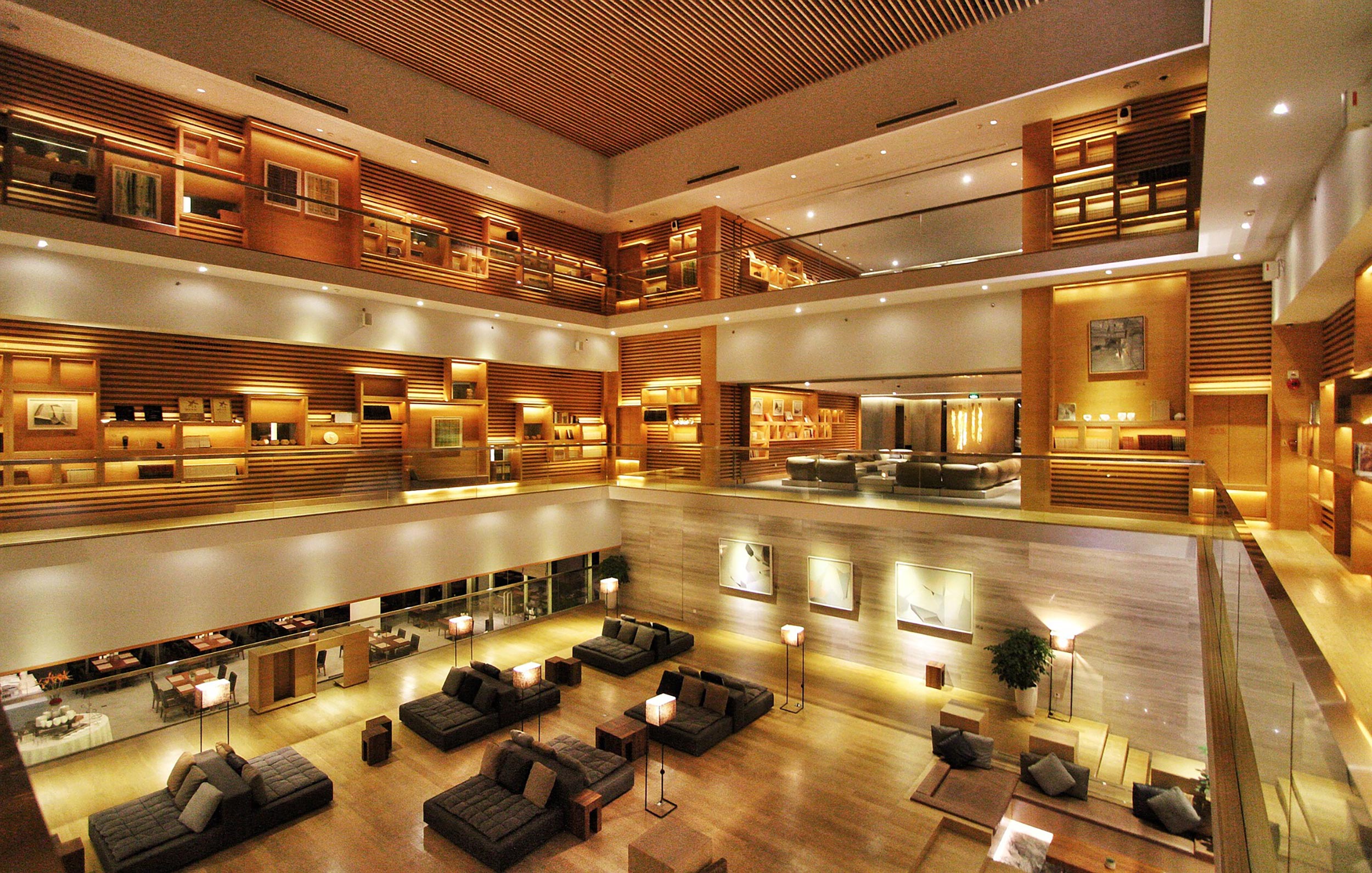 Multi-level hotel atrium featuring warm wood slat walls, illuminated art displays, and clusters of modern lounge seating on the ground floor.