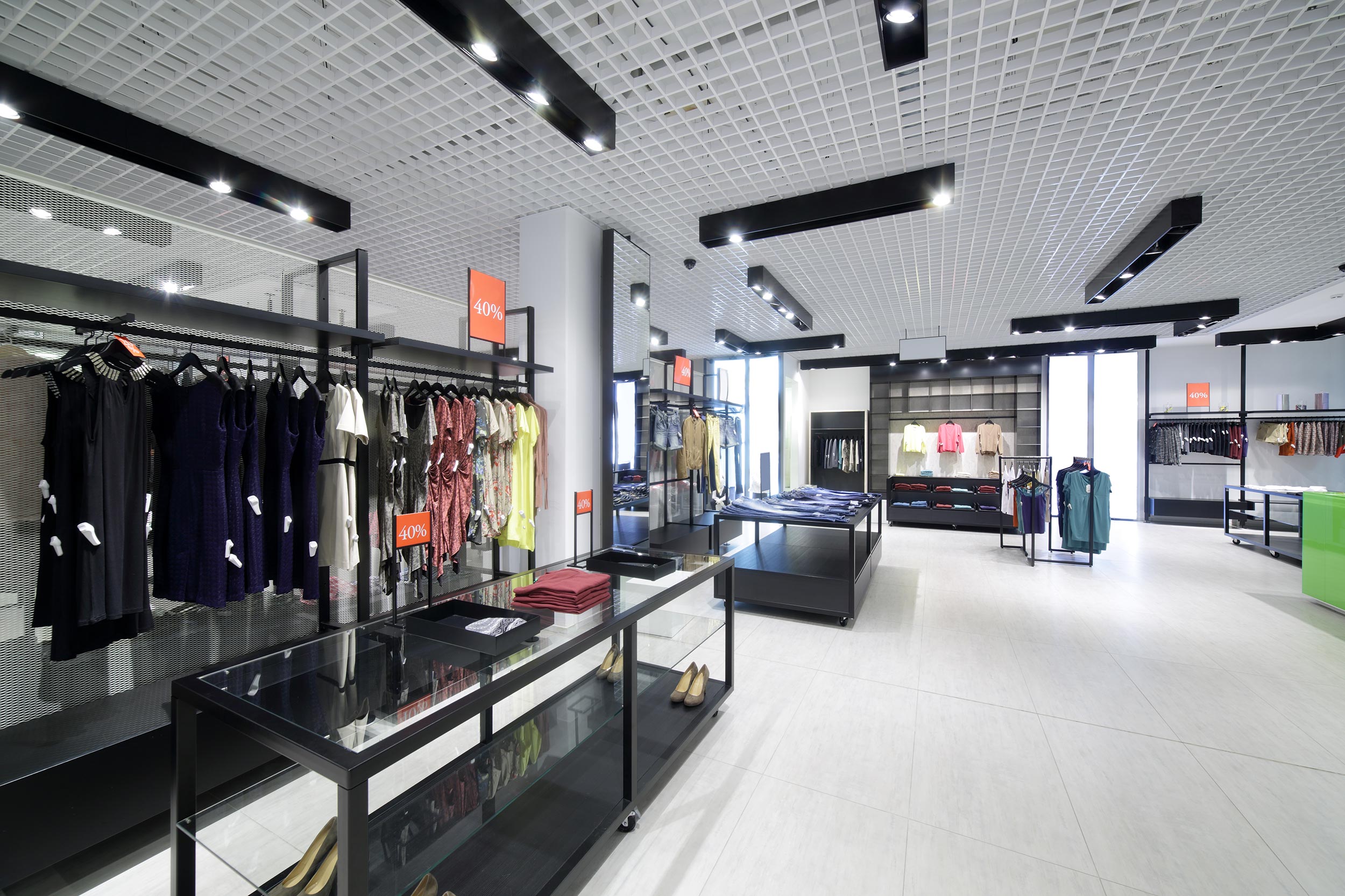 Modern clothing retail store with grid ceiling and track lights, showcasing racks of women’s apparel marked “40 %” off and black metal display tables on a light-tile floor.