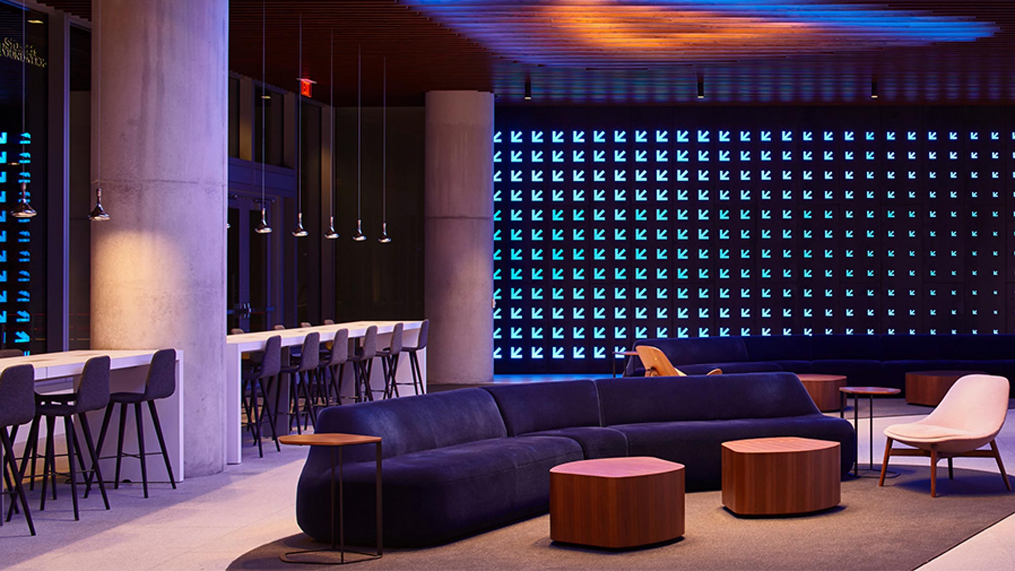 Contemporary lounge with color-changing Ketra lighting: blue arrow-pattern wall and warm amber ceiling wash above modern seating and bar tables.