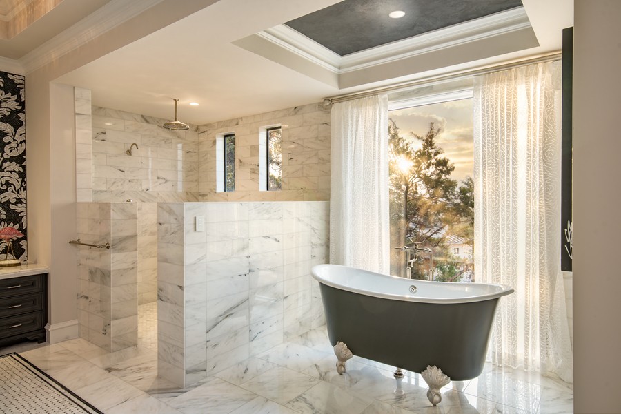 A beautifully illuminated bathroom with a clawfoot bathtub, marble shower, and sunlight entering a window.