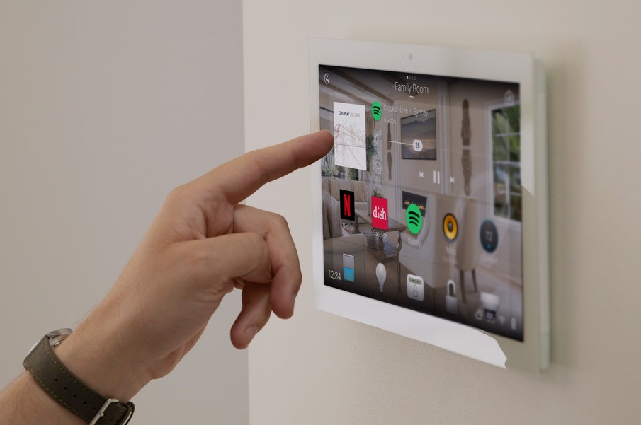 A person's hand using a wall-mounted smart home control panel displaying icons for music, lighting, and security systems.