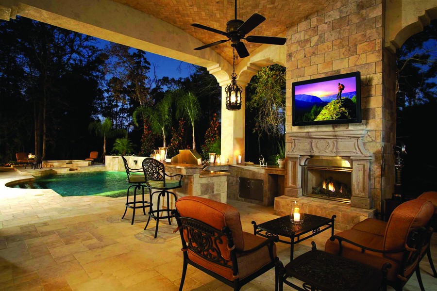 An outdoor patio with seating, a kitchen area, and a Séura outdoor TV above a fireplace.