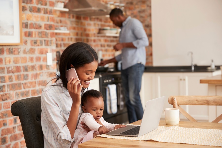 A smiling mother works from home while holding her baby in her lap at a kitchen table, talking on the phone and using a laptop, with a man cooking in the background.