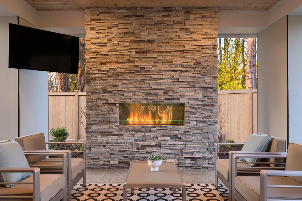 An outdoor living area on a covered patio with a fireplace and a wall-mounted TV on the left wall. 