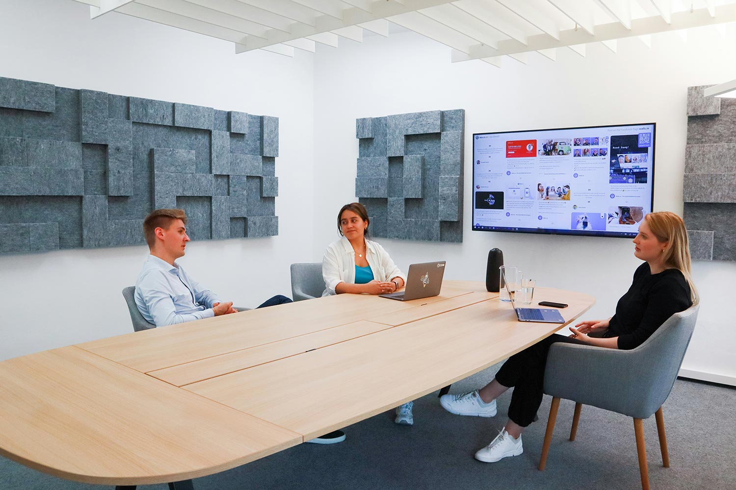 Three people in a modern conference room viewing a wall monitor