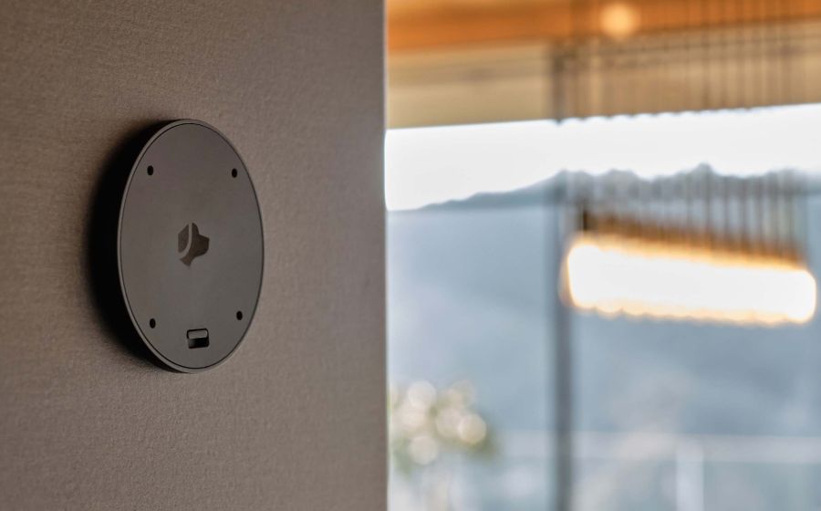 Black circular smart home sensor mounted on an interior wall, with a blurred view of a modern light fixture and large window overlooking hills in the background.