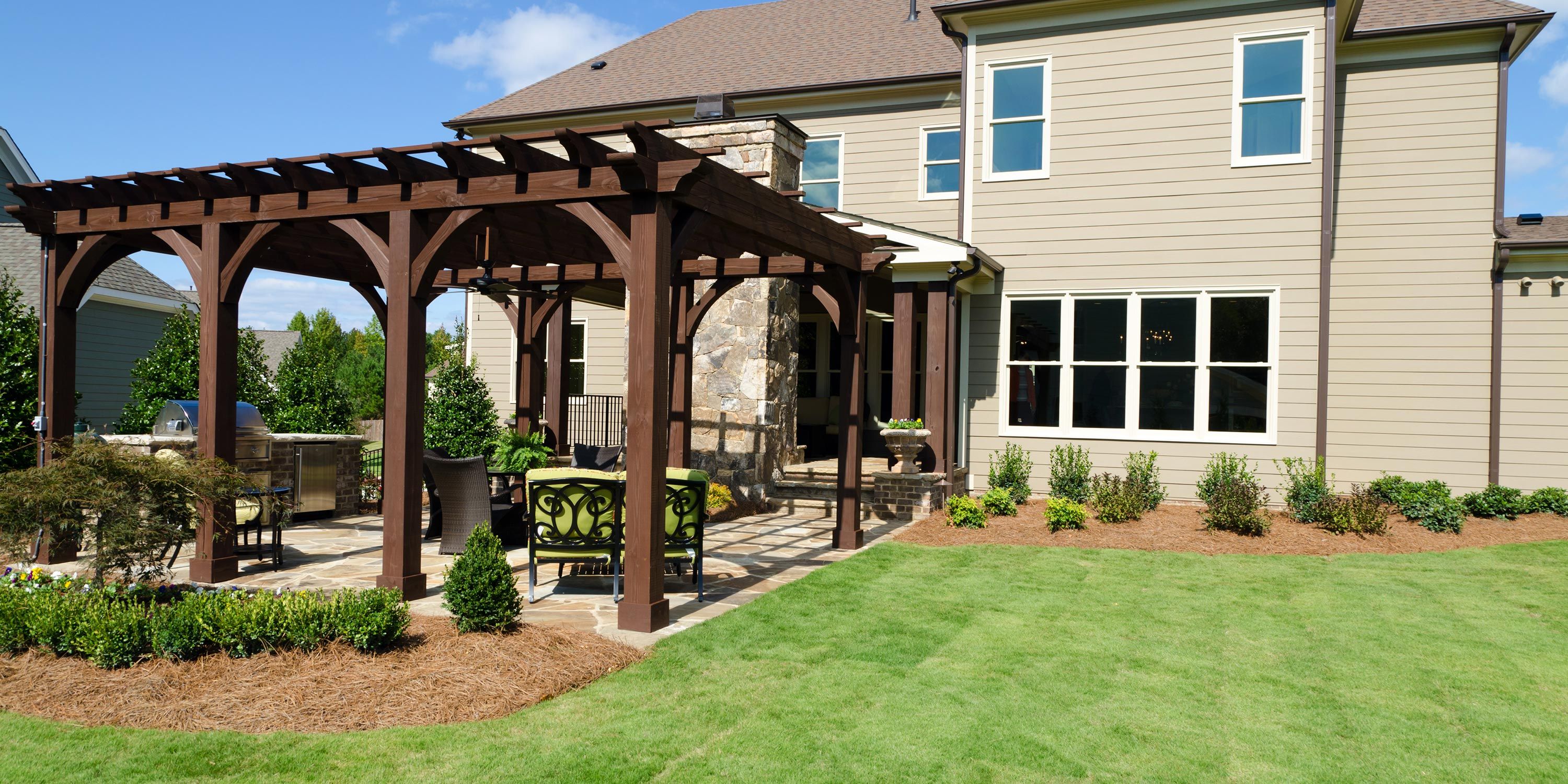 Outdoor kitchen with a wooden pergola, dining set, and well-maintained garden surrounding a stone patio.