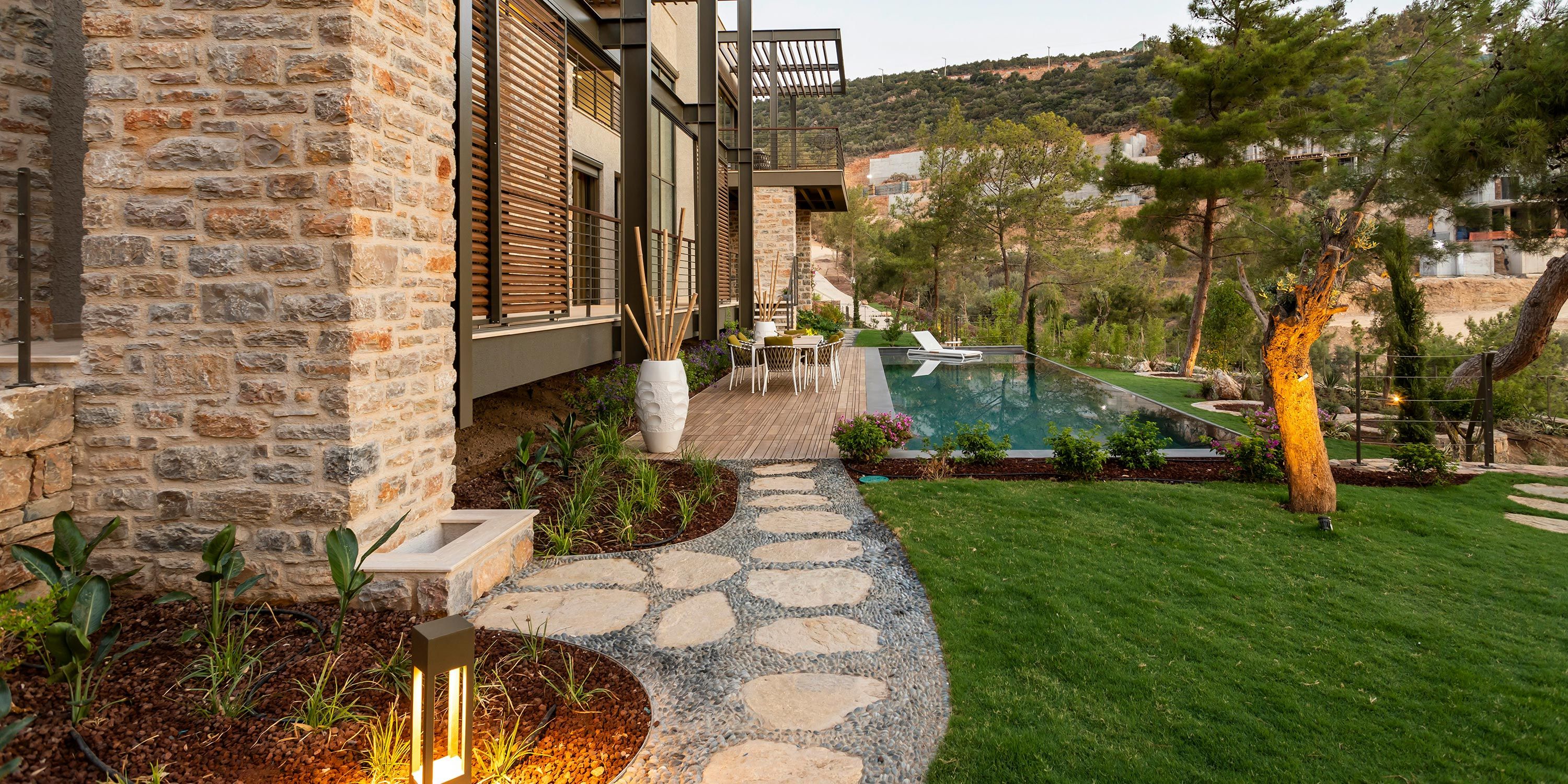 Scenic garden with stone pathway, vibrant plants, and a poolside seating area set against a mountainous backdrop.